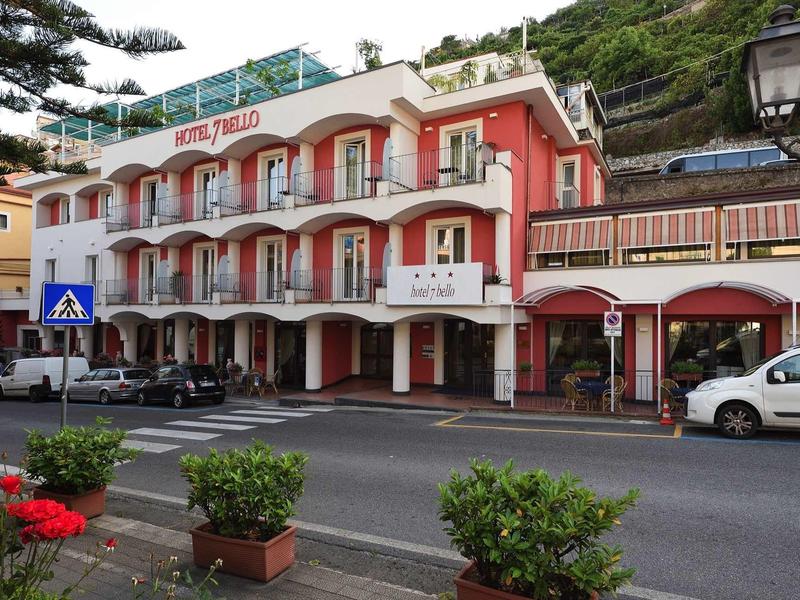 Red hotel building with balconies and terrace on a street with plants and parked cars.