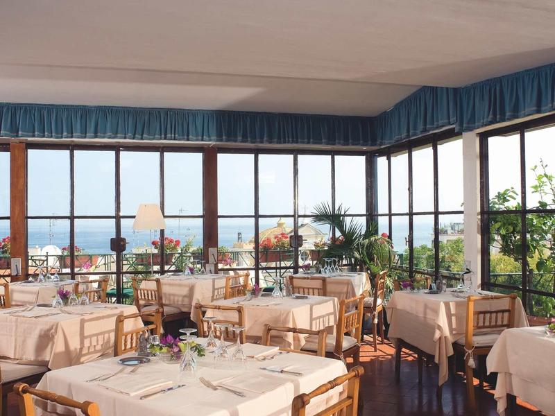 Bright restaurant with large windows overlooking the sea, tables set with white cloths.