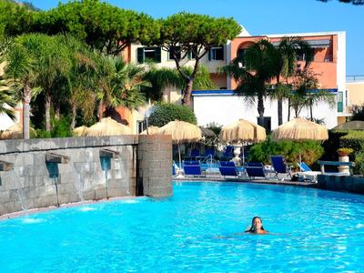 Une grande piscine extérieure avec une eau bleue claire, entourée de chaises longues et de palmiers devant un bâtiment d'hôtel.