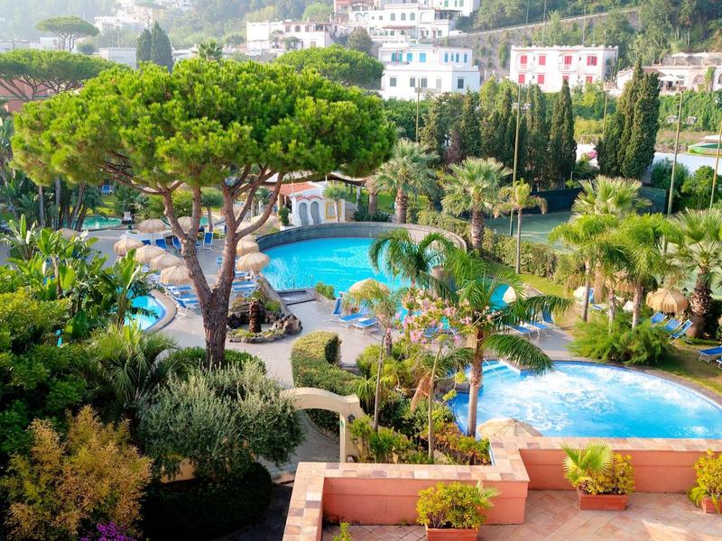 Grand bassin d'hôtel entouré de palmiers et de verdure en plein soleil méditerranéen.