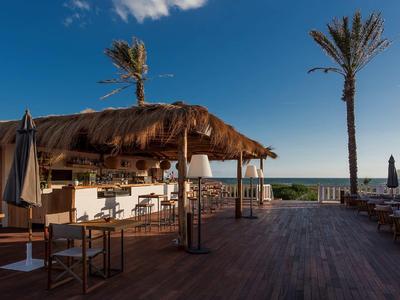 Offene Strandbar mit Palmen, Holzboden und Blick auf das Meer bei klarem Himmel.