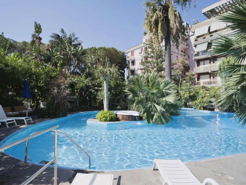 Rond zwembad met ligstoelen en palmbomen voor hotelgebouwen.