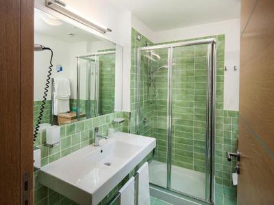 Nowoczesna łazienka z zielonymi płytkami, dużym prysznicem i podwójną umywalką.