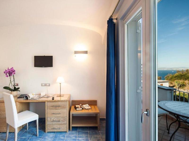 Jasny pokój hotelowy z biurkiem, krzesłem i balkonem z widokiem na morze.