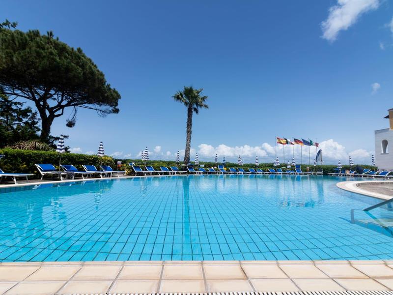 Großer rechteckiger Pool mit blauer Kachelumrandung, Liegestühlen und Palmen bei sonnigem Himmel.