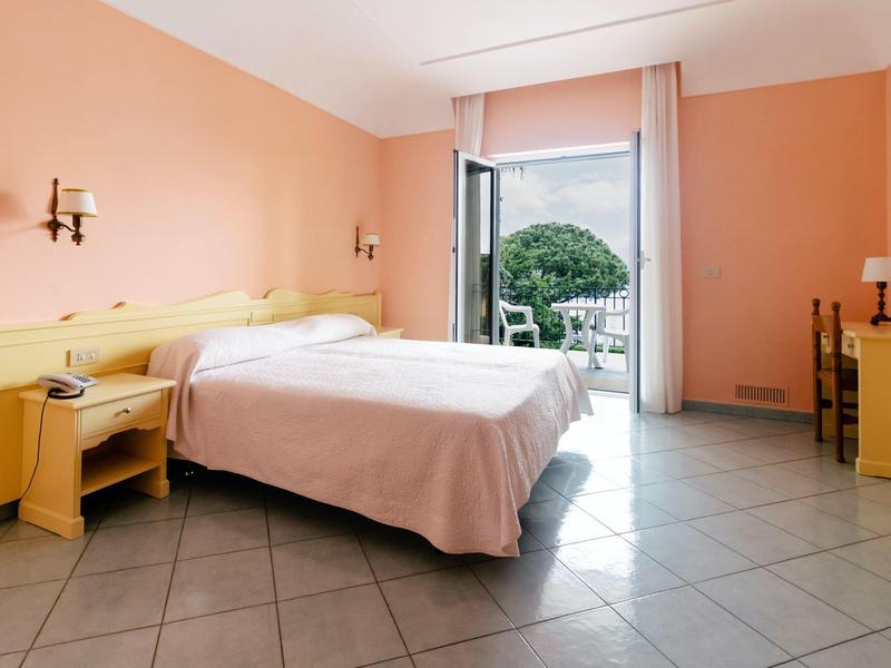 Hell eingerichtetes Schlafzimmer mit Doppelbett, gelben Möbeln und Balkon mit Blick auf Garten.