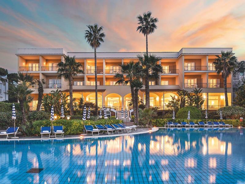 Luxuriöses Hotel mit beleuchteter Fassade, Palmen und großem blauen Pool bei Sonnenuntergang.