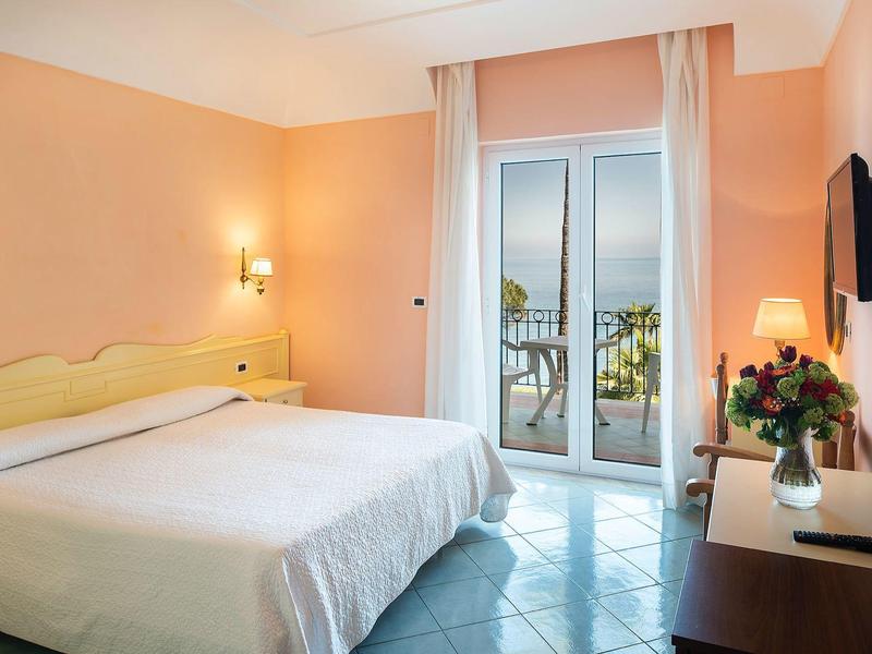 Helles Schlafzimmer mit Doppelbett, Fernseher, Nachttisch und Balkon mit Meerblick.