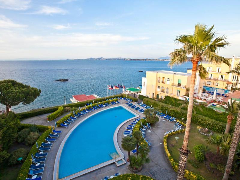 Hotel mit großem Pool, Liegestühlen, Palmen und Blick auf das blaue Meer bei klarem Himmel.
