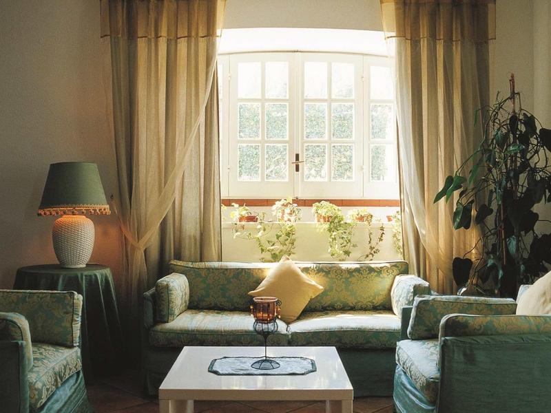 Gemütliches Wohnzimmer mit grünen Sesseln, Sofa, Tisch, Lampen und großen Fenstern mit Vorhängen.