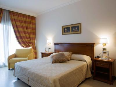 Hell eingerichtet Schlafzimmer mit großem Bett, zwei Nachttischen und gelbem Sessel am Fenster.