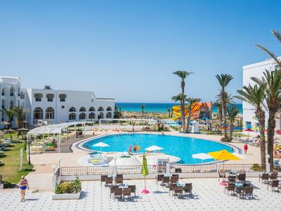 Hotelanlage mit großem Rundpool, Liegestühlen, Palmen und Blick auf das Meer unter klarem Himmel.