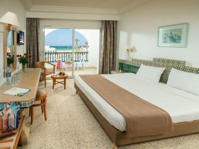 Jasny pokój hotelowy z podwójnym łóżkiem, biurkiem i balkonem z widokiem na morze.