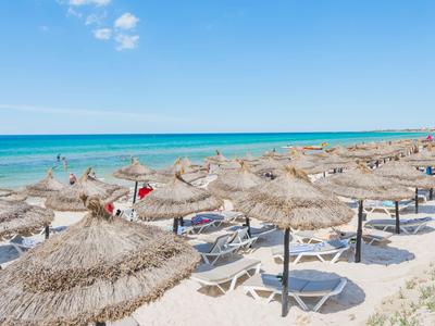 Strand mit vielen Strohhut-Sonnenschirmen und Liegestühlen am Meer bei klarem Himmel.