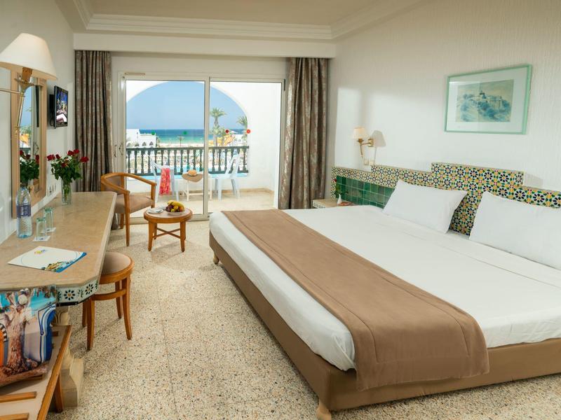 Jasny pokój hotelowy z podwójnym łóżkiem, biurkiem i balkonem z widokiem na morze.