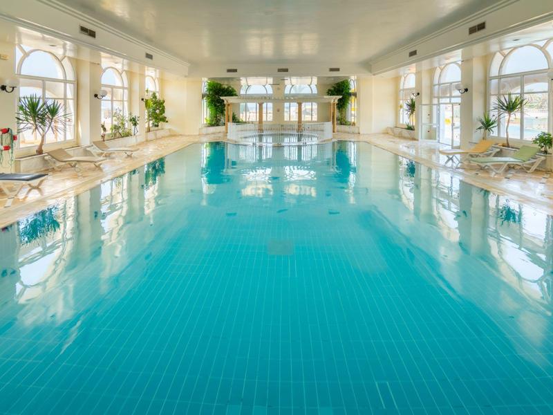 Duży jasny kryty basen z niebieskimi płytkami i roślinami na brzegach.