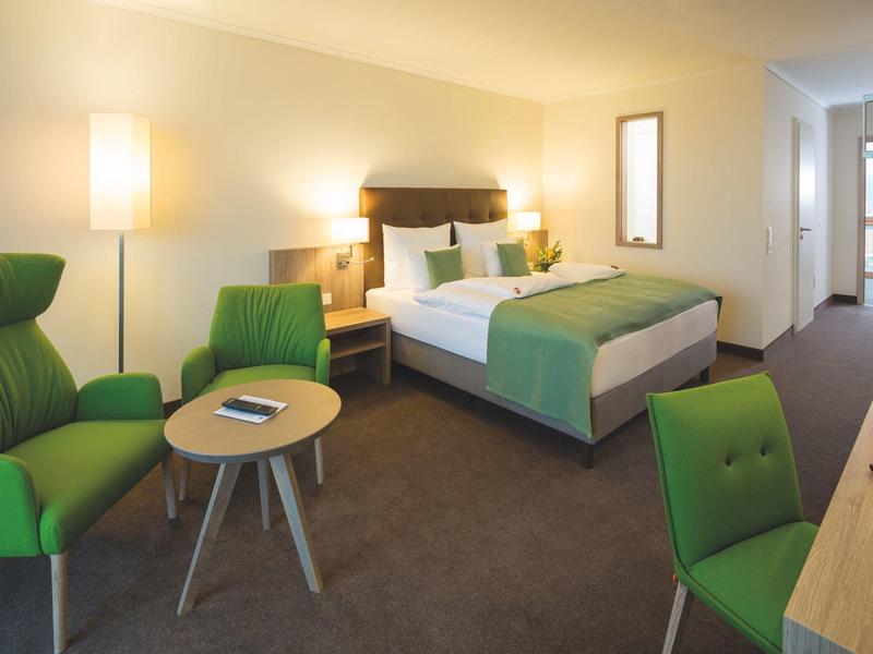 Nowoczesny pokój hotelowy z dużym łóżkiem, zielonymi krzesłami i małym stolikiem z pilotem.