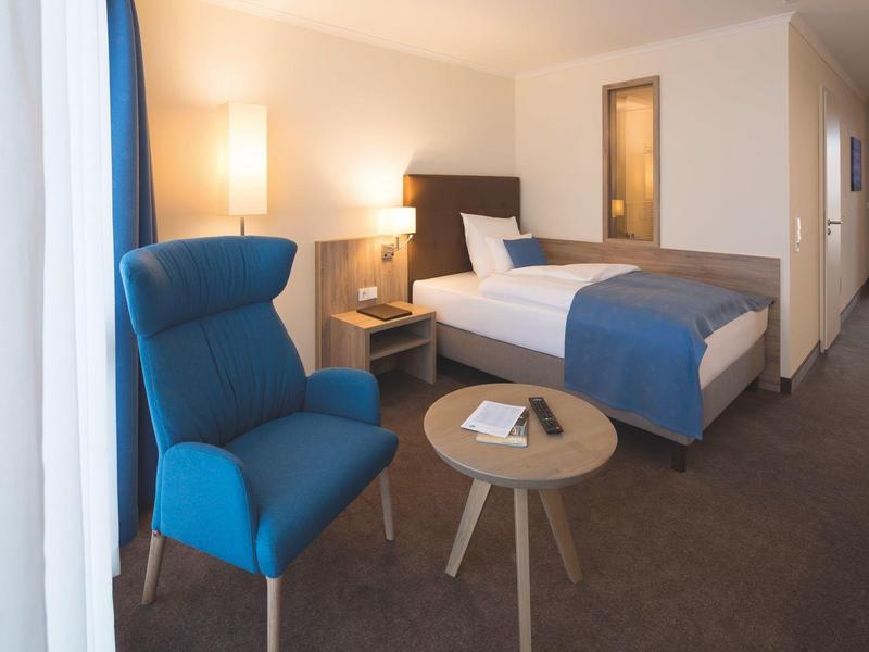 Przytulny pokój hotelowy z niebieskim fotelem, okrągłym stolikiem i dużym łóżkiem w ciepłym świetle.