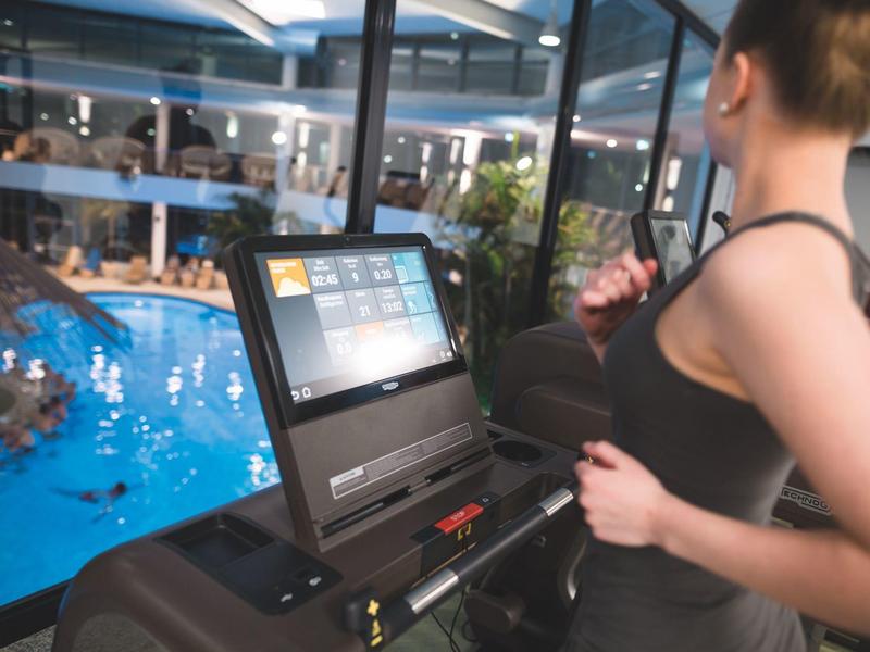Kobieta biegnąca na bieżni z widokiem na kryty basen w hotelowej siłowni.