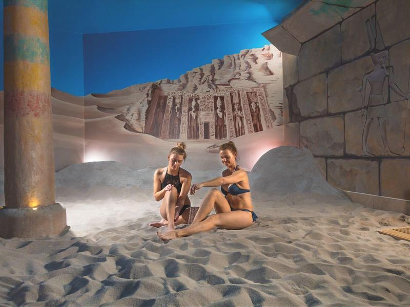 Dwie kobiety w strojach kąpielowych siedzą na piasku w pokoju z egipskimi malowidłami i kolumnami.