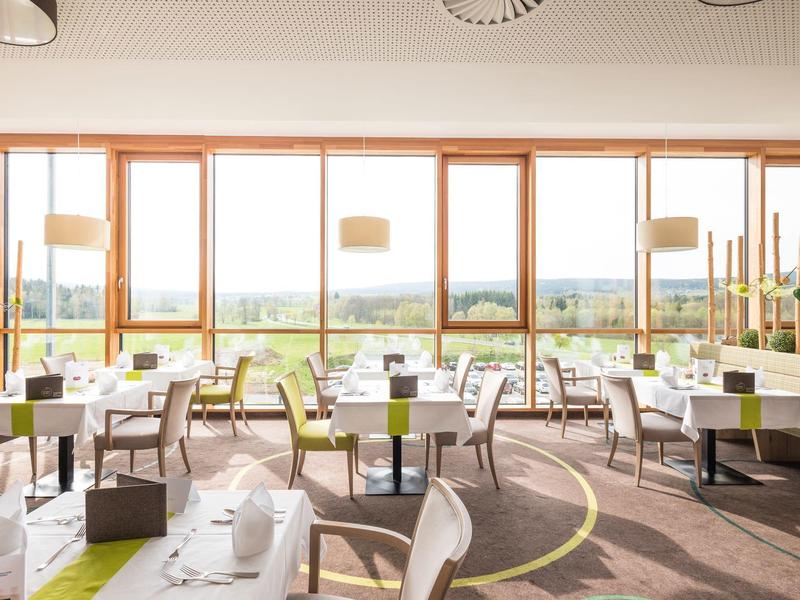 Jasna restauracja z dużymi oknami, białymi stołami i widokiem na krajobraz.