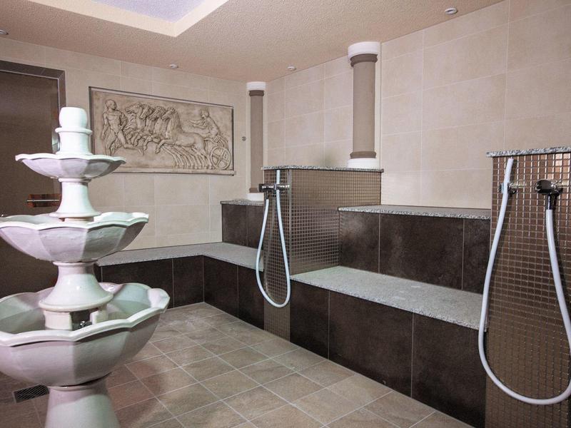 Nowoczesny hammam z marmurową fontanną, dużymi ławkami i prysznicami ręcznymi na ścianach.