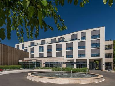 Modernes, weißes Bürogebäude mit Glasfenstern, rundem Springbrunnen und blauem Himmel im Hintergrund.