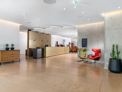 Helle Hotellobby mit Rezeption, modernen Sesseln und grünen Pflanzen in großen Töpfen.