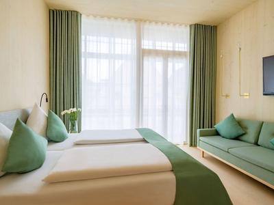 Chambre d'hôtel moderne avec deux lits simples, canapé vert et grande fenêtre avec rideaux.