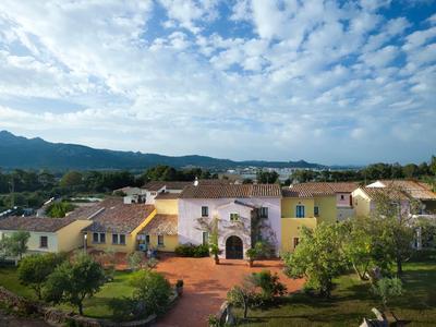 Großes hellgelbes Landhaus mit roten Ziegeldächern, umgeben von Bäumen und Bergen im Hintergrund.