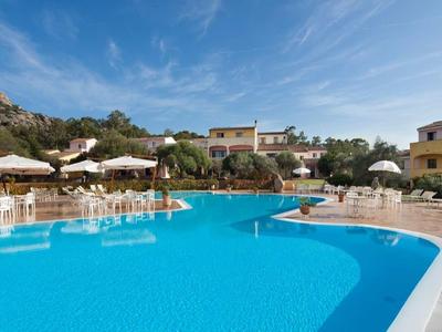 Großer blauer Pool mit Sonnenliegen und Sonnenschirmen umgeben von Gebäuden und grünem Hügel.