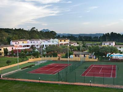 Mehrere rote Tennisplätze mit grünem Zaun, umgeben von Feldern und weißen Gebäuden unter blauem Himmel.