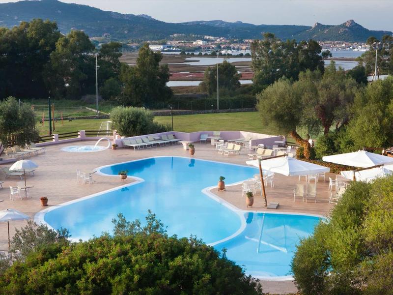 Ein blauer Pool mit weißen Liegestühlen umgeben von grünen Bäumen und Bergen im Hintergrund.