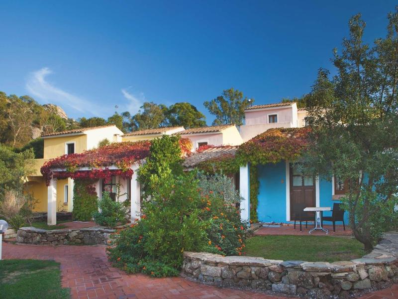 Einfarbiges Haus mit rotem Dach, blauen Türen und üppigen grünen Pflanzen im Garten.