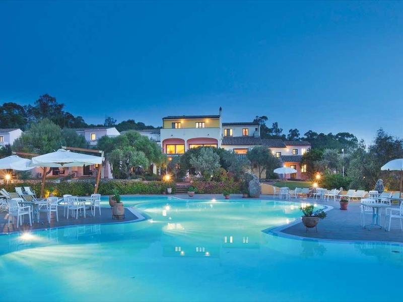 Großer beleuchteter Pool bei Nacht, umgeben von Gebäuden, grünen Hügeln und Sitzbereichen mit Sonnenschirmen.