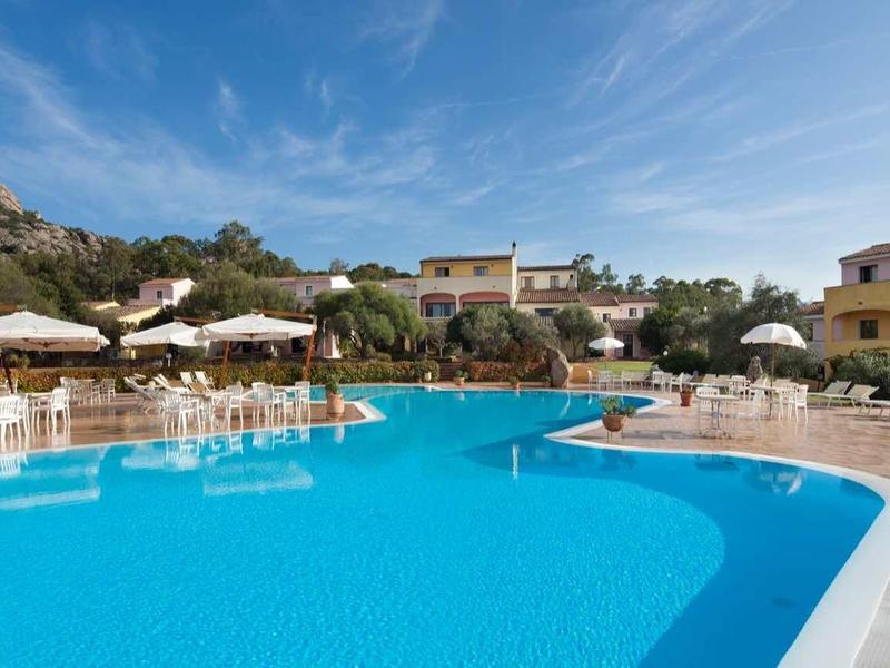 Großer blauer Pool mit Sonnenliegen und Sonnenschirmen umgeben von Gebäuden und grünem Hügel.