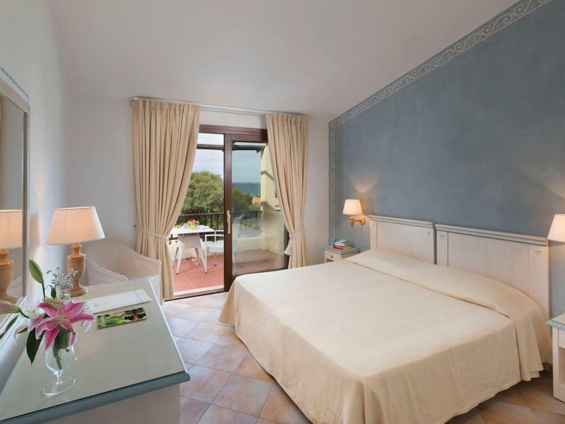 Helles Hotelzimmer mit Doppelbett, Nachttischen, Tisch mit Stuhl und Balkon mit Außensicht.