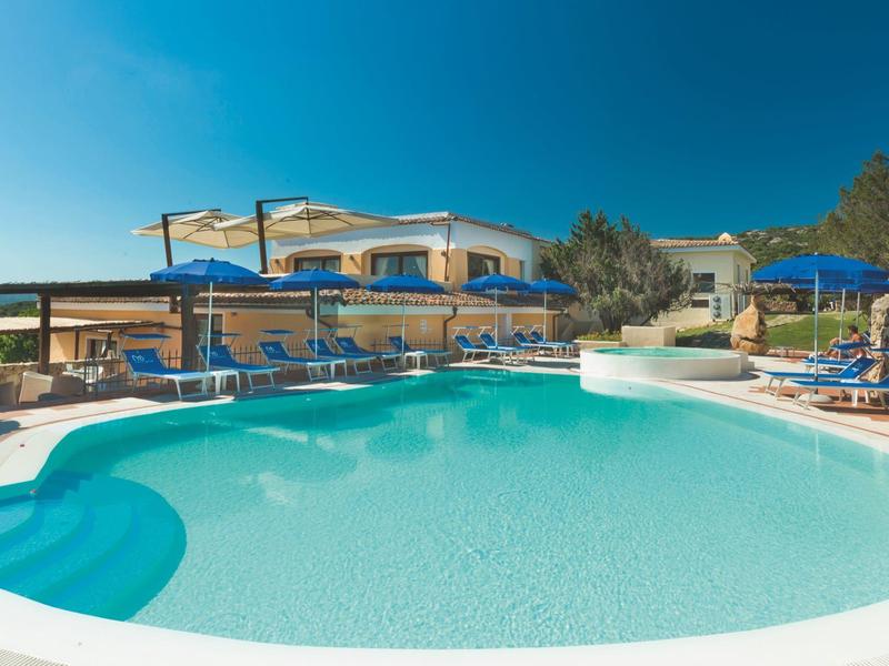 Klares Schwimmbad mit blauen Sonnenschirmen und einer Ferienanlage im Hintergrund