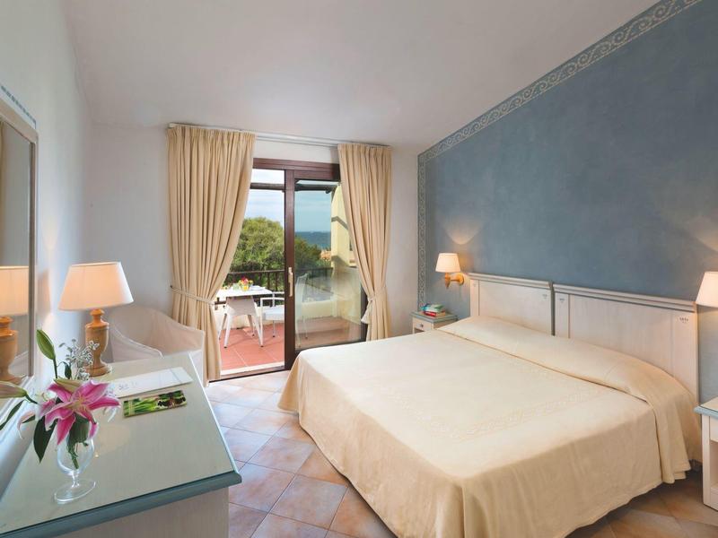 Helles Hotelzimmer mit großem Bett, Sessel, Schreibtisch und Terrassentür mit Blick ins Grüne.