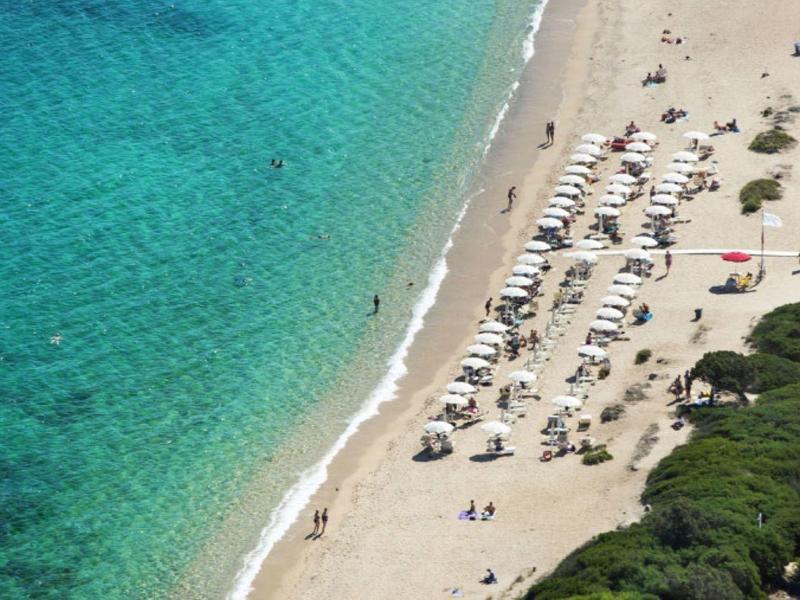 Strand mit türkisfarbenem Wasser, Sand, Sonnenschirmen und Menschen, umgeben von grüner Vegetation.