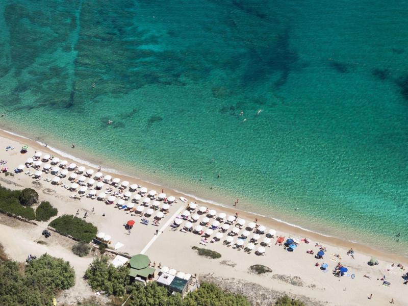 Strand mit weißem Sand, grünen Büschen, Sonnenliegen und klarem türkisfarbenem Wasser.