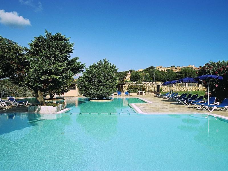 Großer blauer Pool mit Liegestühlen rundherum, Bäumen und klarem Himmel im Hintergrund.
