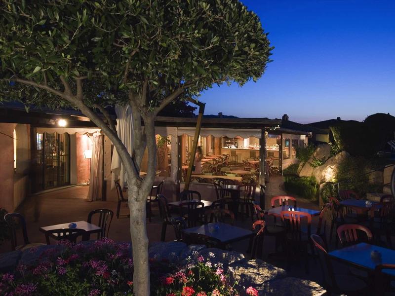 Gemütliche Terrasse mit Tischen, Stühlen, Bäumen und Blumen im Abendlicht unter klarem Himmel.