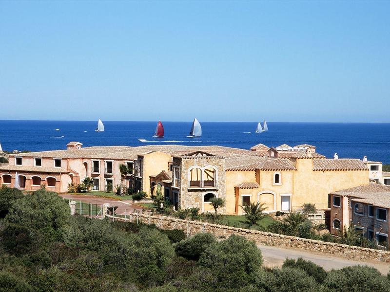 Blick auf mediterrane Ferienhäuser am Meer mit Segelbooten auf dem Wasser.