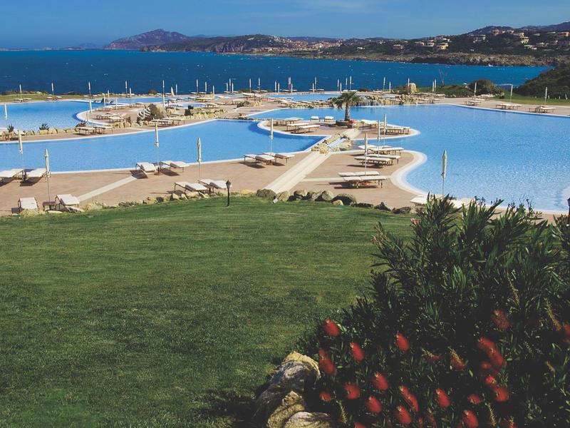 Großer Poolbereich mit Liegestühlen und Meerblick an einem sonnigen Tag.