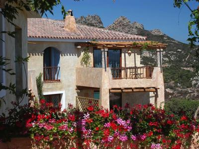 Weißes Haus mit rotem Blumengarten und Bergen im Hintergrund unter blauem Himmel.