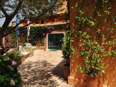 Sonniger Gartenweg mit blühenden Pflanzen, hölzernen Türen und gelb-orangefarbener Hauswand.