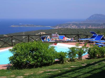 Blick auf einen Pool mit blauen Liegestühlen, Meer im Hintergrund und grünen Sträuchern im Vordergrund.