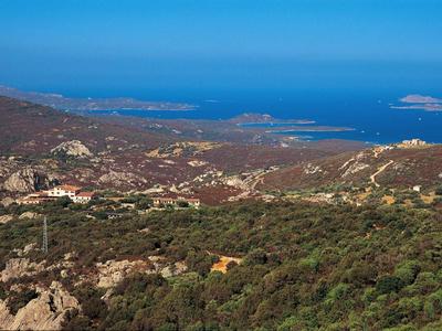 Landschaft mit Hügeln, grünen Büschen, Häusern und blauem Meer im Hintergrund unter klarem Himmel.