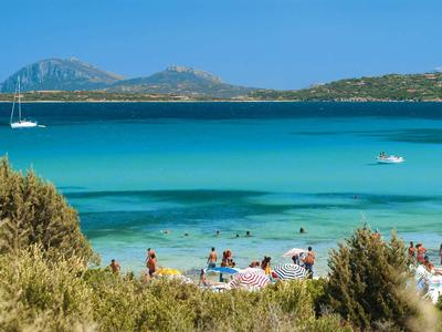 Klares, blaues Meer mit Booten, Sandstrand und Menschen, bewachsene Dünen im Vordergrund.